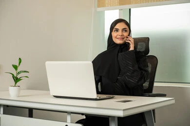 Conducting a business conversation over the phone, browsing social media, completing tasks online, managing and organizing business affairs, a Saudi Arabian Gulf woman wearing a black abaya and using a laptop and mobile phone, lifestyle in upscale companies, gestures of engagement and focus.