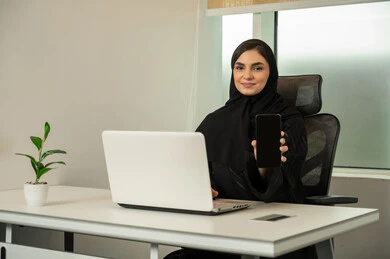 Displaying the mobile phone screen, following global company news, browsing social media, completing tasks online, managing and organizing business affairs, an Arab Gulf Saudi woman wearing a black abaya and using a laptop and mobile phone, lifestyle in a high-end work environment.