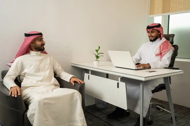 Dialogue and discussion and providing services, friendliness and good reception, answering questions and inquiries, two Arab Gulf men from Saudi Arabia wearing traditional thob and ghutrah sitting in the office, with one of them using a laptop, a comfortable work environment in upscale companies.