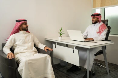Answering questions and inquiries, dialogue and discussion, and providing services, friendliness and good reception, two Arab Gulf Saudi men wearing traditional thob and ghutrah sitting in the office, with one of them using a laptop, a comfortable work environment in upscale companies.