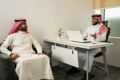 Data collection and experience exchange, friendliness and good reception, answering questions and inquiries, two Arab Gulf men from Saudi Arabia wearing traditional thobes and ghutras sitting in the office, with one of them using a laptop, a comfortable work environment in upscale companies.