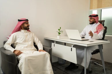 Providing services to customers, friendliness and good reception, answering questions and inquiries, two Arab Gulf Saudi men wearing traditional thob and ghutrah sitting in the office, with one of them using a laptop, a comfortable work environment in upscale companies, discussion and dialogue among colleagues.