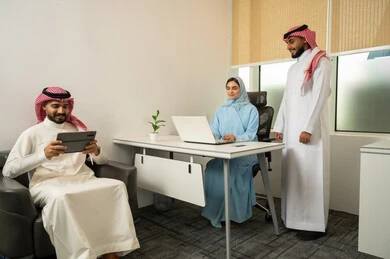 تبادل الخبرات و المعلومات لتحقيق المنفعة العامة,  بيئة عمل السعودية, فريق عربي خليجي سعودي مكون من رجلين يرتديان الثوب التقليدي و الشماغ و سيدة محجبة ترتدي العباءة الزرقاء و أمامها جهاز اللابتوب, دمج الأعمال مع التقنية و التكنولوجيا, البشاشة و حسن الاستقبال