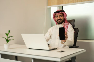 النظر الى الكاميرا أثناء عرض شاشة المحمول, استعمال الحاسب و التقنية في مجال الاعمال، مفهوم التركيز و الدقة في الإنجاز، مهنة و وظيفة سعودية مكتبية, إتمام مهام العمل في المكتب، موظف عربي خليجي سعودي يرتدي الثوب التقليدي و الشماغ و يقوم باستخدام اللابتوب والموبايل