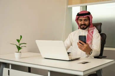 Completing tasks via mobile phone, using computers and technology in the field of business, the concept of focus, accuracy, and mastery, a Saudi office profession and job, completing work tasks in the office, an Arab Gulf Saudi employee wearing traditional attire and a headscarf while using a laptop and mobile phone.