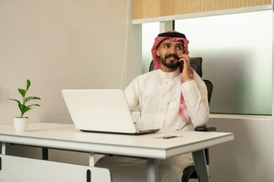 Making a phone call, using computers and technology in the field of business, the concept of focus and accuracy in achievement, a Saudi office profession and job, completing work tasks in the office, an Arab Gulf Saudi employee wearing traditional attire and a headscarf while using a laptop and a mobile phone.