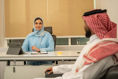 Reviewing meeting results, organizing and preparing daily work reports, business feasibility studies, administrative professions and jobs, teamwork and collaboration, a Saudi Gulf Arab team consisting of a man wearing traditional attire and a ghutrah and a woman in a hijab wearing a blue abaya with a tablet in front of her.