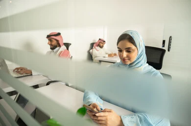 Business and company development, work meetings and project completion, discussing plans and explaining work tasks, Arab Saudi Gulf colleagues wearing traditional attire and shemagh, and a lady wearing a blue abaya using modern devices, designated spaces for each employee in the company.