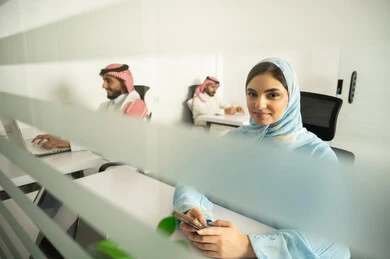 Focus and meticulous work, dialogues and discussions within the team, business and company development, work meetings and project completion, discussing plans and explaining work tasks, Arab Saudi Gulf colleagues wearing traditional attire and shemagh, and a woman wearing a blue abaya using modern devices.