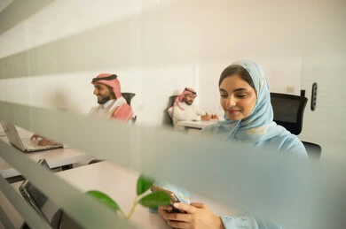 Designated spaces for each employee in the company, focus and meticulous work, dialogues and discussions within the team, business and company development, work meetings and project completion, discussing plans and explaining work tasks, Arab Saudi Gulf colleagues wearing traditional attire and shemagh using modern devices.