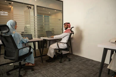 Solving common challenges, teamwork to achieve goals, lifestyle in Saudi companies, an Arab Gulf Saudi team consisting of a man wearing traditional dress and a shemagh and a woman in a hijab wearing a blue abaya, accomplishing tasks through a laptop and tablet.