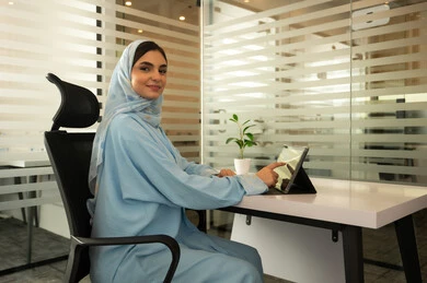 استخدام تطبيقات و برامج الأجهزة اللوحية، استعمال التكنولوجيا و التقنيات الحديثة، الدردشة مع الأهل و الأصدقاء،  سيدة عربية خليجية سعودية مبتسمة محجبة ترتدي عباءة زرقاء و أمامها تابلت,  مهن و وظائف مكتبية