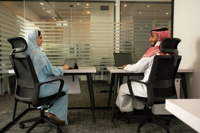 Teamwork to achieve goals, lifestyle in Saudi companies, a Gulf Arab Saudi team consisting of a man wearing traditional attire and a shemagh and a woman in a hijab wearing a blue abaya, accomplishing tasks through a laptop and a tablet.
