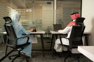 Working on solutions for daily problems, the spirit of collaboration and public conversations, strategic plans to achieve goals, the lifestyle in Saudi companies, a Gulf Arab Saudi team consisting of a man wearing traditional dress and a headscarf and a woman in a hijab wearing a blue abaya, completing tasks using a laptop and tablet.
