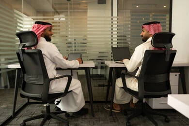 Organized professional environment, working on solutions for daily problems, collaborative spirit and public discussions, strategic plans to achieve goals, lifestyle in Saudi companies, two Arab Gulf Saudi men wearing traditional thob and ghutrah sitting in the office and using a laptop and tablet.