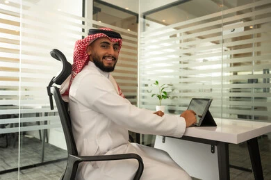 Performing the daily task inside the company headquarters, looking at the camera with expressions of happiness and joy, an Arab Gulf Saudi employee wearing traditional attire and a headscarf, using a tablet, integrating technology and modern devices with practical life.