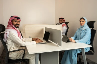 إتمام مهام العمل في المكتب، تطور الشركات السعودية, العمل الجماعي و التعاون، فريق عربي خليجي سعودي مكون من رجلين يرتديان الثوب التقليدي و الشماغ و سيدة محجبة ترتدي العباءة الزرقاء و يقومون باستخدام اللابتوب و التابلت,  النظر الى الكاميرا بتعبيرات السعادة و السرور
