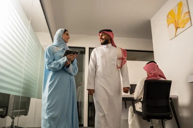 نقاش و حوار في بيئة مكتبية, استخدام الجهاز اللوحي لتوضيح العمليات المتبعة، التفاهم والتعاون بين الزملاء لتحقيق أهداف الشركة، مهن ووظائف إدارية، فريق عربي خليجي سعودي مكون من رجل يرتدي الثوب التقليدي و الشماغ و سيدة محجبة ترتدي العباءة الزرقاء و تشرح استيراتيجية العمل لزميلها باستعمال التابلت