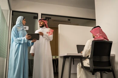 بيئة العمل المريحة في الشركات السعودية، استخدام الحاسب و التقنية في مجال الاعمال، الحوار و النقاش بين الزملاء، تبادل الخبرات و المعلومات لتحقيق الاهداف,  فريق عربي خليجي سعودي مكون من رجل يرتدي الثوب التقليدي و الشماغ و سيدة محجبة ترتدي العباءة الزرقاء و تشرح استيراتيجية العمل لزميلها باستخدام التابلت