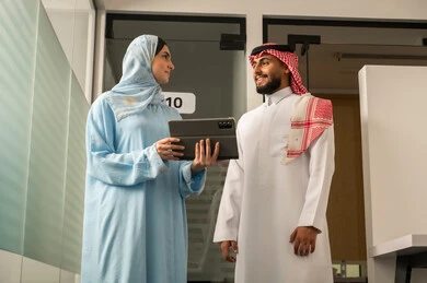 Dialogue and understanding among employees, unity and teamwork, professions and office jobs, a Saudi Gulf Arab team consisting of a man wearing traditional attire and a shemagh and a hijab-wearing woman in a blue abaya explaining the strategy to her colleague using a tablet, ensuring a clear and comprehensive understanding of the work.