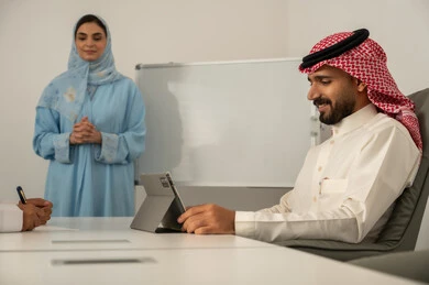 استخدام التابلت في تدوين الملاحظات، استعمال السبورة لتوضيح العمليات و الاجراءات المتبعة في الشركة، العمل الجماعي و التعاون، فريق عربي خليجي سعودي مكون من رجل يرتدي الثوب التقليدي و الشماغ و سيدة محجبة ترتدي العباءة الزرقاء و تشرح الاستيراتيجية لزميلها, ضمان فهم واضح و شامل للاعمال