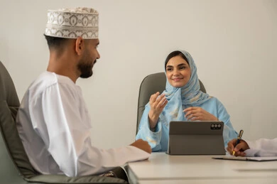 شرح المهام المطلوبة بسعادة وحب, ورشات عمل في مقر الشركة، استخدام أجهزة تقنية حديثة، جمع البيانات وحل المشكلات، اجتماع عمل بين رجل عربي خليجي عماني يرتدي الدشداشة و الكمة و سيدة محجبة ترتدي عباءة زرقاء و تستخدم التابلت, الحوارات و النقاشات حول المشاريع