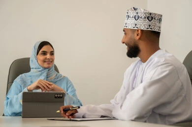 Management and organization of the company's work, use of modern technological devices, data collection and problem-solving, a business meeting between an Arab Gulf Omani man wearing a dishdasha and kumma and a hijab-wearing woman in a blue abaya using a tablet, dialogues and discussions about projects, collaboration and participation and exchange of opinions, writing and taking notes.