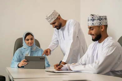Collaboration and participation in completing tasks, new partnership deals, dialogues and discussions about projects, developing plans and achieving the company's goals, a Gulf Arab Omani team consisting of two men wearing the dishdasha and kumma and a veiled woman wearing a blue abaya, pointing with the index finger at the tablet, writing and taking notes.