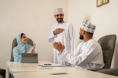 Cooperation and love among employees, developing plans and achieving the company's goals, collaboration and participation in completing tasks, a Gulf Arab Omani team consisting of a man wearing a dishdasha and a kumma and a hijab-wearing woman in a blue abaya in front of a tablet, clapping and congratulating their colleague.