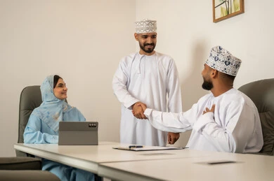صفقات شراكة جديدة، الحوارات و النقاشات حول المشاريع، تطوير الخطط وتحقيق أهداف الشركة، التعاون و المشاركة في إنجاز الأعمال،  فريق عمل عربي خليجي عماني مكون من رجلين يرتديان الدشداشة و الكمة و سيدة محجبة ترتدي عباءة زرقاء, مصافحة و سلام حار بين الزملاء