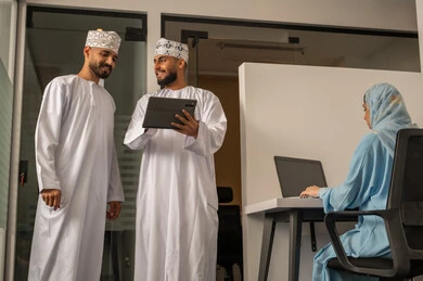 تقسيم المهام بين الموظفين, متابعة المهام اليومية، إنجاز الأعمال المكتبية، الحوارات و النقاشات حول المشاريع، تطوير الخطط وتحقيق أهداف الشركة، فريق عمل عربي خليجي عماني مكون من رجلين يرتديان الدشداشة و الكمة يشير أحدهما الى التابلت و سيدة محجبة ترتدي عباءة زرقاء تجلس على المكتب و تستخدم اللابتوب