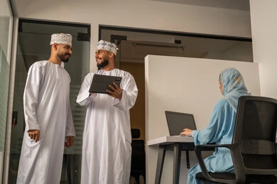 مراقبة سير المهام اليومية، إنجاز الأعمال المكتبية، الحوارات و النقاشات حول المشاريع، تطوير الخطط وتحقيق أهداف الشركة، فريق عمل عربي خليجي عماني مكون من رجلين يرتديان الدشداشة و الكمة يحمل أحدهما التابلت و سيدة محجبة ترتدي عباءة زرقاء تجلس على المكتب و تستخدم اللابتوب