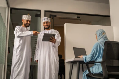 Monitoring daily tasks, completing office work, discussions and dialogues about projects, developing plans and achieving the company's goals, a Gulf Arab Omani team consisting of two men wearing the dishdasha and kumma, one of whom is pointing to the tablet, and a veiled woman wearing a blue abaya sitting at the desk and using a laptop.