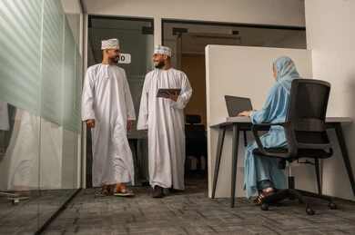 Discussion of work methods, success and achieving goals, the concept of dialogue and exchanging opinions, monitoring the progress of daily tasks, completing office work, an Arab Gulf Omani team consisting of two men wearing dishdasha and kumma, one of whom is holding a tablet and walking, and a veiled woman wearing a blue abaya sitting at the desk and using a laptop.