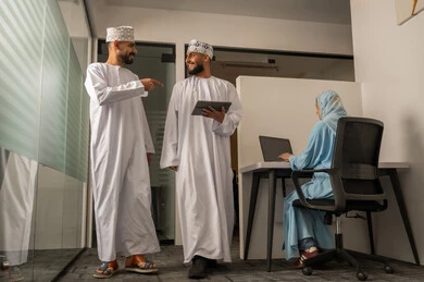 النجاح و تحقيق الأهداف, مفهوم التحاور و تبادل الآراء, مراقبة سير المهام اليومية، إنجاز الأعمال المكتبية، فريق عمل عربي خليجي عماني مكون من رجلين يرتديان الدشداشة و الكمة يحمل أحدهما تابلت و يقومان بالمشي و سيدة محجبة ترتدي عباءة زرقاء تجلس على المكتب و تستخدم اللابتوب