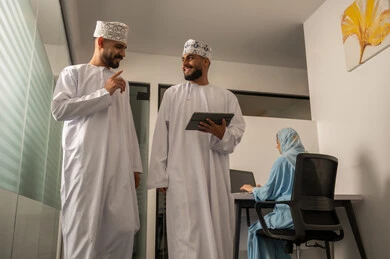 Exchanging conversations among colleagues, monitoring the progress of daily tasks, completing office work, developing plans and achieving the company's goals, a Gulf Arab Omani team consisting of two men wearing dishdasha and kumma, one of whom is holding a tablet, and a veiled woman wearing a blue abaya sitting at the desk and using a laptop.