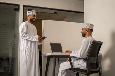 تعليم الموظفين الأساليب الجديدة في العمل لتحقيق أهداف الشركة، دمج الأعمال مع التقنية و التكنولوجيا، رجلان عربيان خليجيان عمانيان مبتسمان يرتديان الدشداشة و الكمة يتناقشان  في المكتب, انجاز المهام من خلال الحاسوب المحمول,  بيئة العمل في سلطنة عمان, مفهوم التعاون و روح الفريق