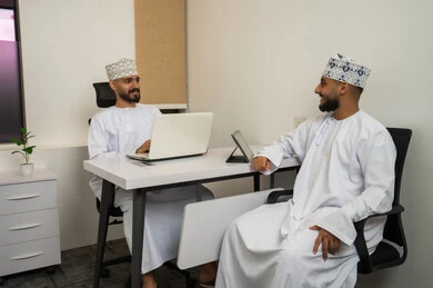 Teamwork and the spirit of one team, discussing proposed deals, the work environment in the Sultanate of Oman, expressions and gestures of happiness and joy, two smiling Arab Gulf Omani men wearing the dishdasha and kumma sitting in the office and discussing, completing tasks through the laptop and tablet.