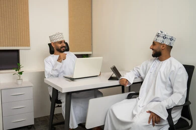 Discussion of proposed deals, the work environment in the Sultanate of Oman, the concept of cooperation and joint work, expressions and gestures of happiness and joy, two smiling Arab Gulf Omani men wearing the dishdasha and kumma sitting in the office and discussing, accomplishing tasks through the laptop and tablet.