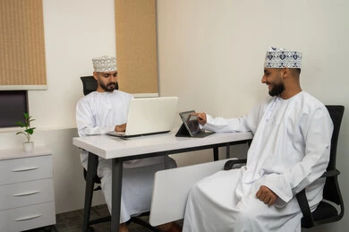 إدارة العمليات و تنظيم الأعمال، تحليل البيانات و إيجاد الحلول، التعاون و روح الفريق الواحد, بيئة العمل في سلطنة عمان, تعابير وايماءات السعادة والسرور، رجلان عربيان خليجيان عمانيان مبتسمان يرتديان الدشداشة و الكمة يجلسان في المكتب و يتناقشان, انجاز المهام من خلال الحاسوب المحمول و التابلت