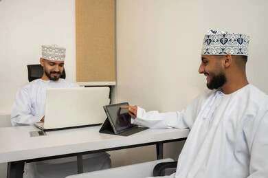 تحليل البيانات و إيجاد الحلول، التعاون و روح الفريق الواحد, بيئة العمل في سلطنة عمان, تعابير وايماءات السعادة والسرور، رجلان عربيان خليجيان عمانيان مبتسمان يرتديان الدشداشة و الكمة يجلسان في المكتب و يتناقشان, انجاز المهام من خلال الحاسوب المحمول و التابلت