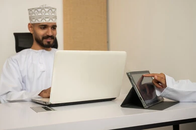 A comfortable professional environment in the institutions of the Sultanate of Oman, accomplishing tasks through a laptop, expressions of happiness and joy, an Arab Gulf Omani man wearing a dishdasha and kumma using a laptop, entering data through the keyboard, integrating technology and modern devices with practical life.