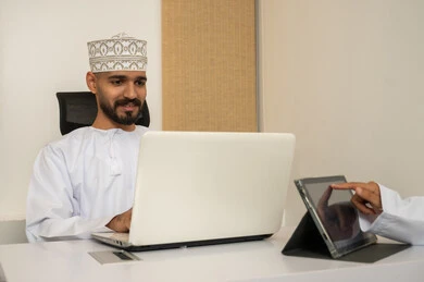 Professionalism and dedication to work, accomplishing tasks through a laptop, a prestigious Omani company, expressions of happiness and joy, an Arab Gulf Omani man wearing a dishdasha and kumma using a laptop, data entry through the keyboard, integrating technology and modern devices with practical life.