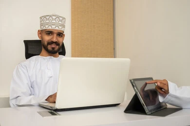 Completing tasks using a laptop, an Omani work environment, looking at the camera with expressions of happiness and joy, an Arab Gulf Omani man wearing a dishdasha and kumma using a laptop, entering data through the keyboard, integrating technology and modern devices with practical life.