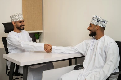 التعارف و بناء العلاقات الاجتماعية، مفهوم الترحيب و السلام و إلقاء التحية، بيئة العمل المريحة في الشركات الراقية، مفهوم التعاون والعمل المشترك, تعابير وايماءات السعادة والسرور، رجلان عربيان خليجيان عمانيان مبتسمان يرتديان الدشداشة و الكمة يجلسان في المكتب و يقومان بمصافحة بعضهم, استخدام الأجهزة المتطورة
