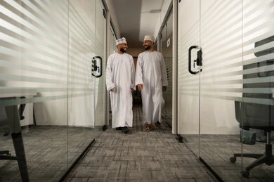 تبادل اطراف الحديث, جودة الحياة في الشركات الراقية, الفن الهندسي المعماري لمكاتب الأعمال، المشي في ممر الشركة بين الابواب الزجاجية الحديثة, مهن مكتبية إدارية, رجلان عربيان خليجيان عمانيان يرتديان الدشداشة و الكمة يتمشيان