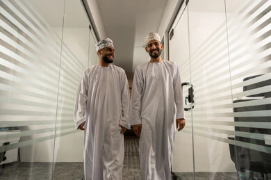 جودة الحياة في الشركات الراقية,  الفن الهندسي المعماري لمكاتب الأعمال، المشي في ممر الشركة بين الابواب الزجاجية الحديثة, مهن مكتبية إدارية, رجلان عربيان خليجيان عمانيان يرتديان الدشداشة و الكمة يتمشيان و يتبادلان أطراف الحديث