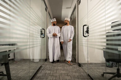 Walking in the company corridor, modern contemporary design for corporate offices in the Sultanate of Oman, professions and administrative office work, two Arab Gulf Omani men wearing the dishdasha and kumma are walking and exchanging conversation.