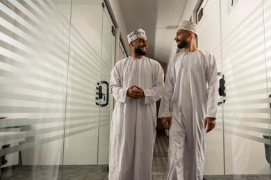 الفن الهندسي المعماري لمكاتب الأعمال، المشي في ممر الشركة بين الابواب الزجاجية الحديثة, تصميم عصري حديث لمكاتب الشركات في سلطنة عمان, مهن مكتبية إدارية, رجلان عربيان خليجيان عمانيان يرتديان الدشداشة و الكمة يتمشيان و يتبادلان أطراف الحديث