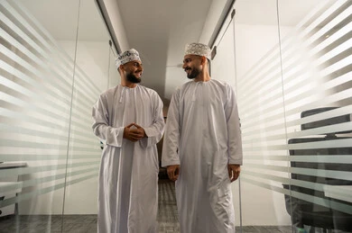 Walking in the company corridor between the modern glass doors, a modern contemporary design for corporate offices in the Sultanate of Oman, administrative office professions and jobs, two Arab Gulf Omani men wearing the dishdasha and the kumma are walking and exchanging conversation.